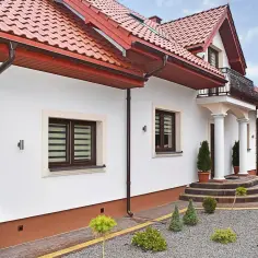 Elegancki dom z białym tynkiem i brązowymi detalami. Cokół wykończony ozdobnym tynkiem mozaikowym dodaje mu charakteru.