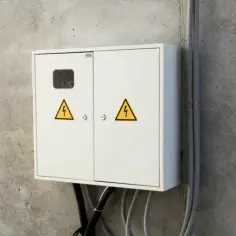 Biała skrzynka elektryczna na betonowej ścianie, z żółtymi symbolami ostrzegawczymi. To miejsce, gdzie skrzynka elektryczna na działce zapewnia zasilanie.