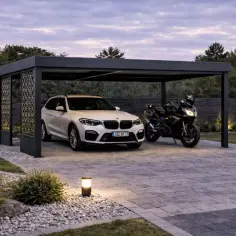 Nowoczesna wiata garażowa ażurowa chroni białe BMW i czarny motocykl. Oświetlony ogród i kamienna nawierzchnia tworzą elegancką całość.