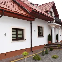 Elegancki dom z białymi ścianami i brązowymi rynnami. Wokół domu zadbany ogród z kamyczkami i iglakami.