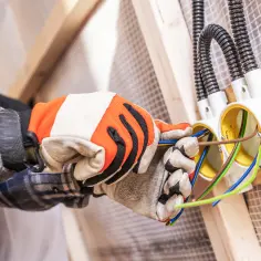 Ręce w rękawicach montują puszki elektryczne, przygotowując instalacje elektryczne w domu. Widać przewody w kolorach zielonym, niebieskim i brązowym.