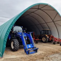 Zielona wiata bez pozwolenia wymiary chroni niebieski traktor New Holland i żółty traktor z maszynami rolniczymi.