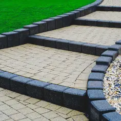 Eleganckie schody wejściowe do domu, wyłożone kostką brukową, otoczone ciemnymi kamieniami i zielonym trawnikiem.