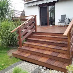 Wysoki taras ze schodami prowadzi do drzwi tarasowych. Drewniana konstrukcja otoczona zielenią.