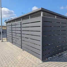 Nowoczesne wiaty śmietnikowe w Warszawie, wykonane z ciemnoszarego metalu, z kratowanymi elementami dachu.