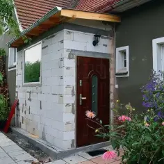 Nowoczesny wiatrołap zewnętrzny w trakcie budowy, z białej cegły, z brązowymi drzwiami i oknem. Obok kwitnące kwiaty.