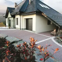 Nowoczesny podjazd przed domem z kamienia i kostki brukowej, otoczony zielenią i skalnymi elementami.