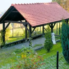Drewniana wiata z czerwonym dachem, idealna na boisko do gry w bule. Otoczona zielenią i jesiennymi drzewami.