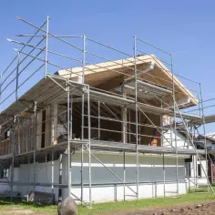 Budowa dobudówki do starego domu. Widać rusztowania, drewnianą konstrukcję dachu i białe ściany.