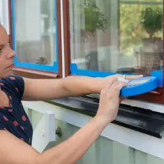 Kobieta okleja taśmą malarską okno, przygotowując je do malowania. To prosty sposób, jak zrobić okno na nowo.