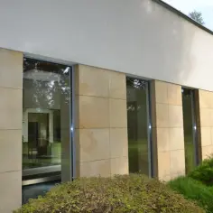 Nowoczesna elewacja z piaskowca, z dużymi oknami odbijającymi zieleń.