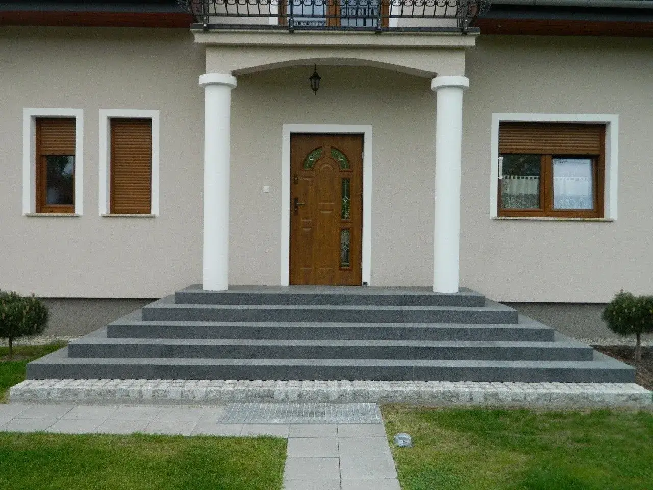 Szerokie schody wejściowe do domu prowadzą do brązowych drzwi. Dwa białe filary podtrzymują daszek nad wejściem.