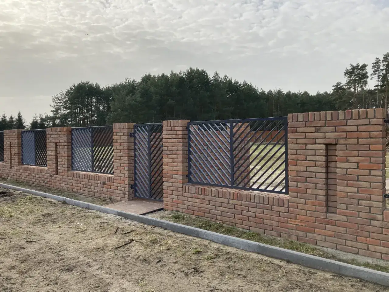 Solidny ogrodzenie z cegły pełnej z metalowymi wstawkami i bramą, otaczające teren z widokiem na las.
