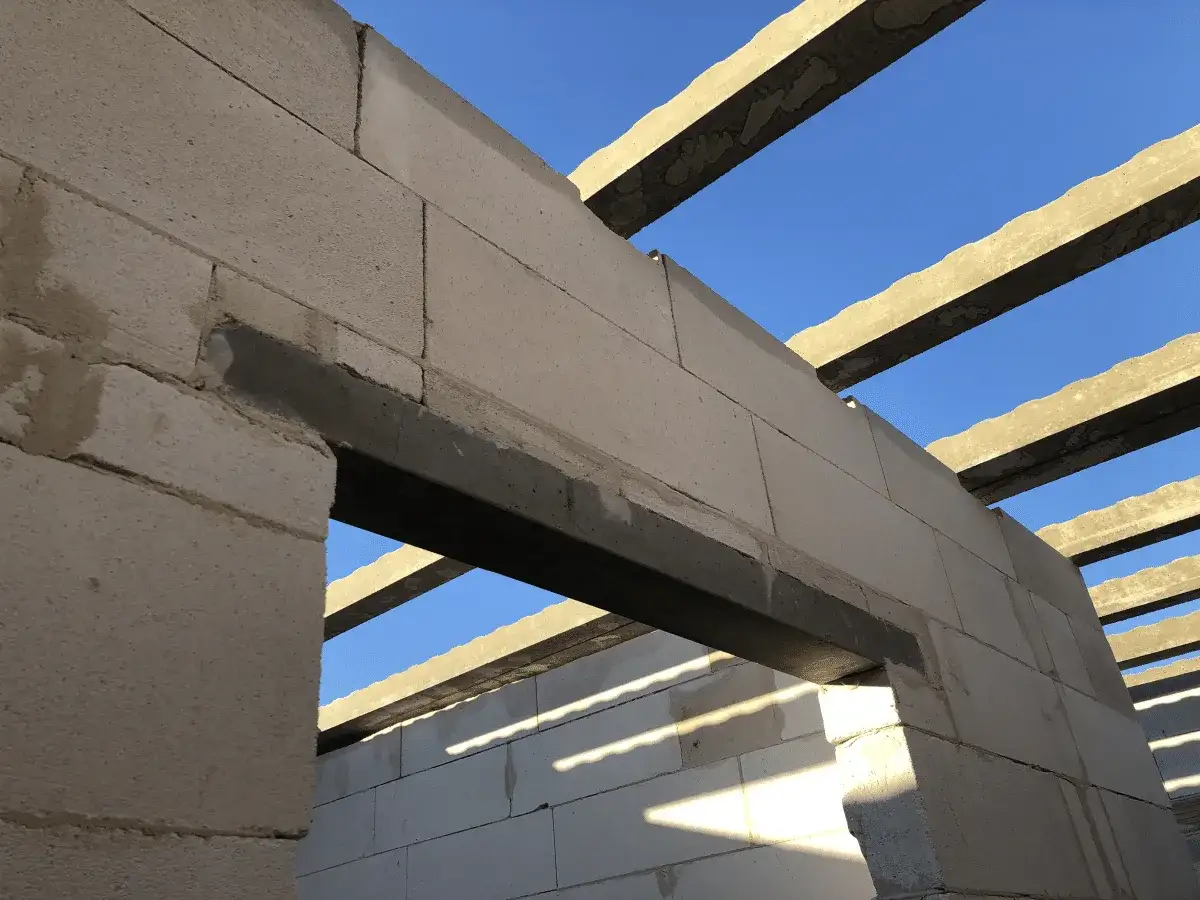 Betonowe nadproże nad otworem okiennym w budowie, widoczne belki stropowe i błękitne niebo.