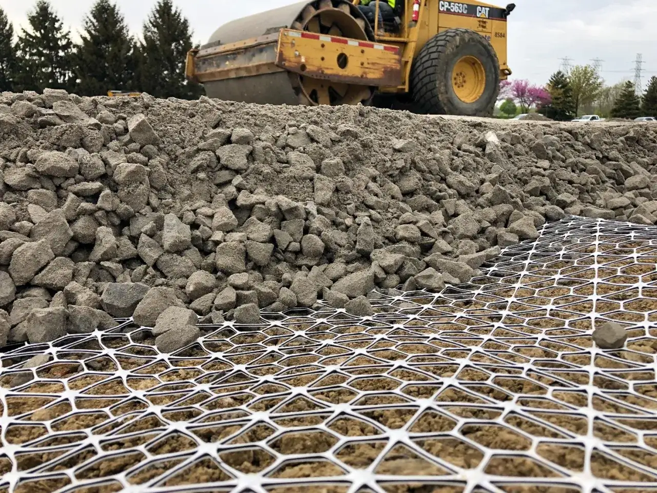 Na placu budowy walec drogowy ugniata kruszywo do betonu. Pod nim widoczna jest biała siatka geotechniczna.