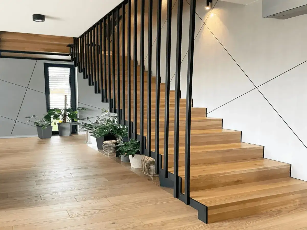 Nowoczesne schody w domu jednorodzinnym z drewnianymi stopniami i czarną, metalową balustradą. Wnętrze rozjaśniają rośliny doniczkowe.
