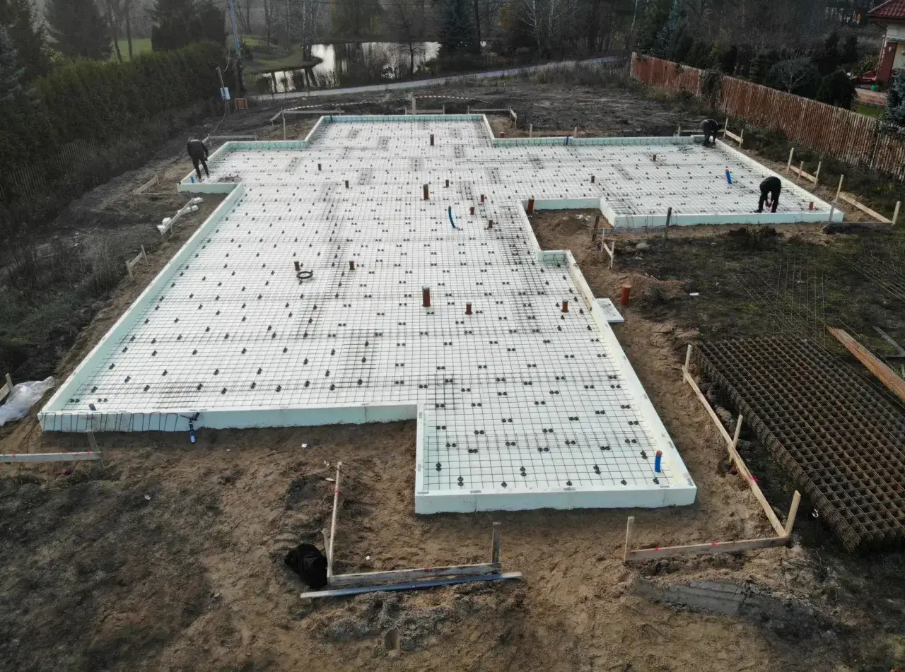 Przygotowanie wylewki betonowej pod garaż blaszany. Na placu budowy widać zbrojenie i izolację fundamentów.