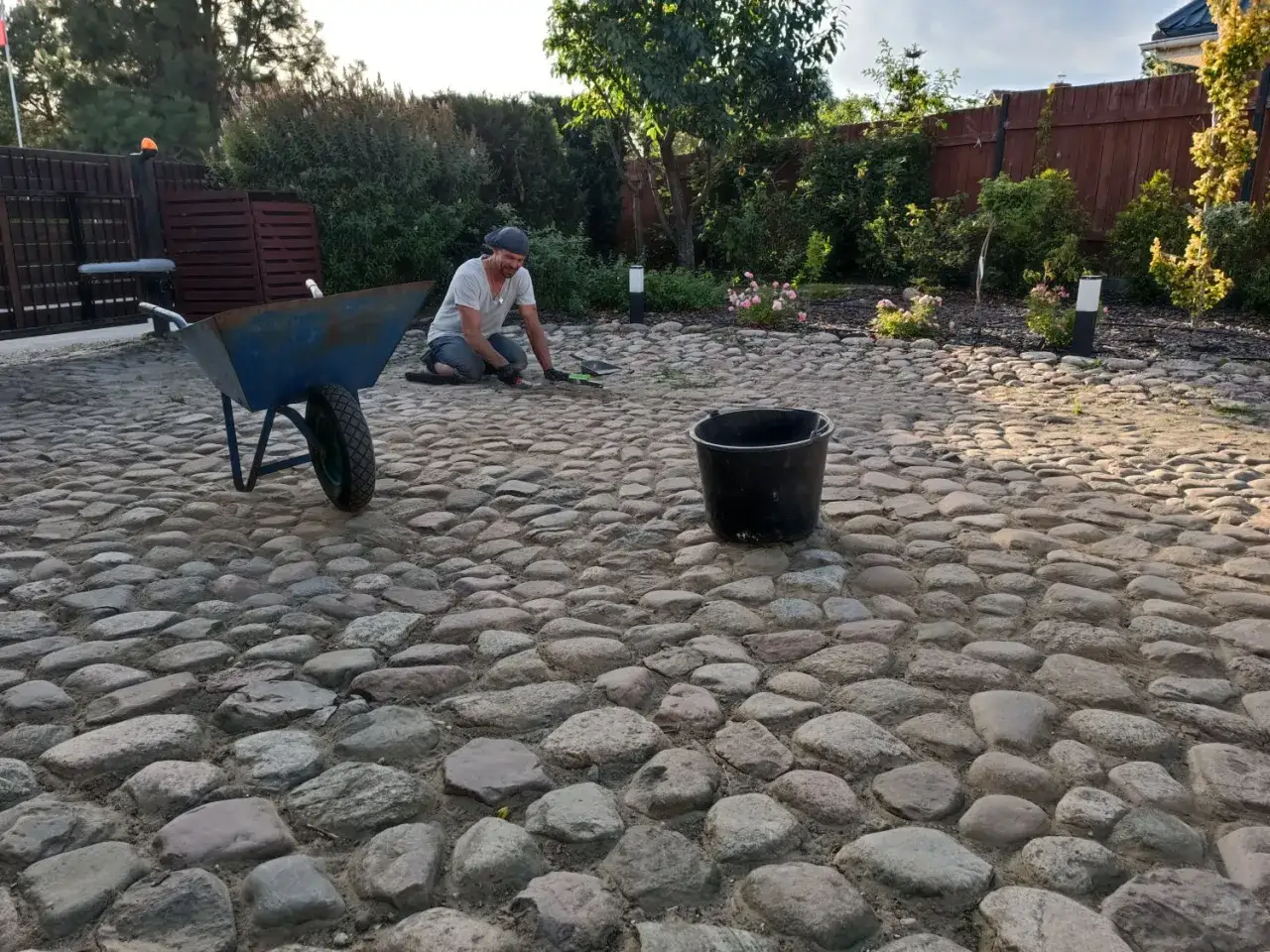 Mężczyzna pracuje przy budowie podjazdu z kamienia polnego. Obok stoi taczka i wiadro.