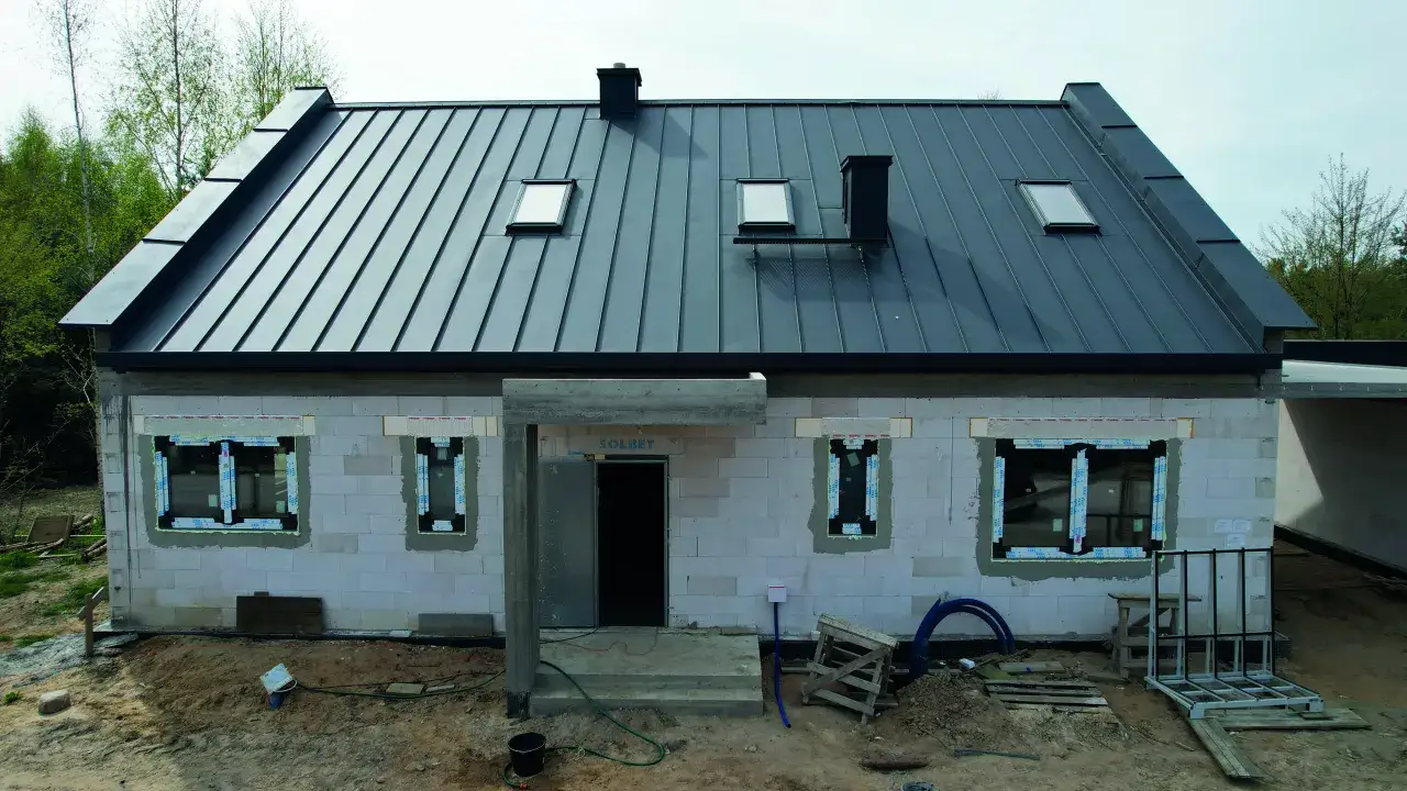 Nowy dom w budowie. Widoczny styk ściany szczytowej z dachem pokrytym czarną blachą. Widać okna dachowe i komin.