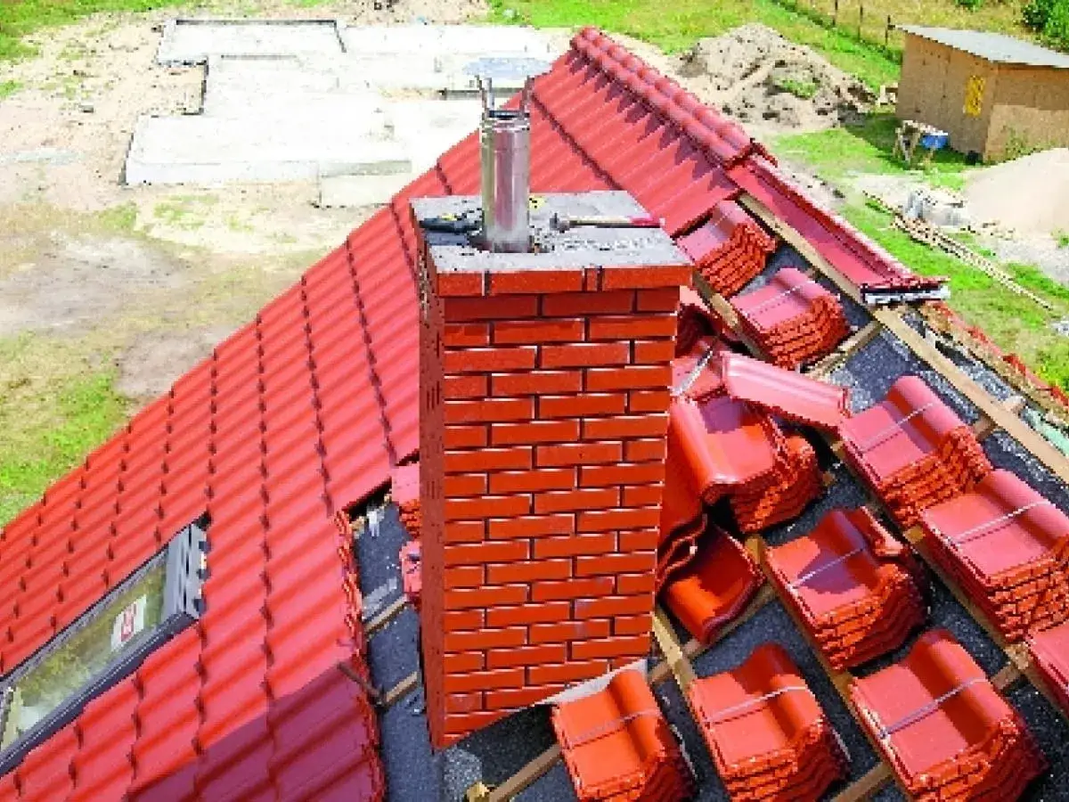Ceglany komin na dachu z czerwonej dachówki, obok stosy dachówek czekających na ułożenie.