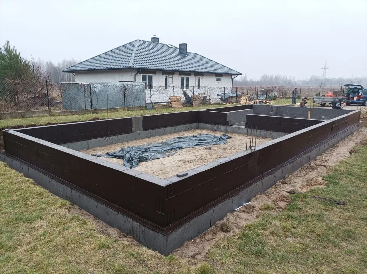 Fundament domu w budowie, ciemnobrązowy cokol wokol domu, w tle dom i plac budowy.