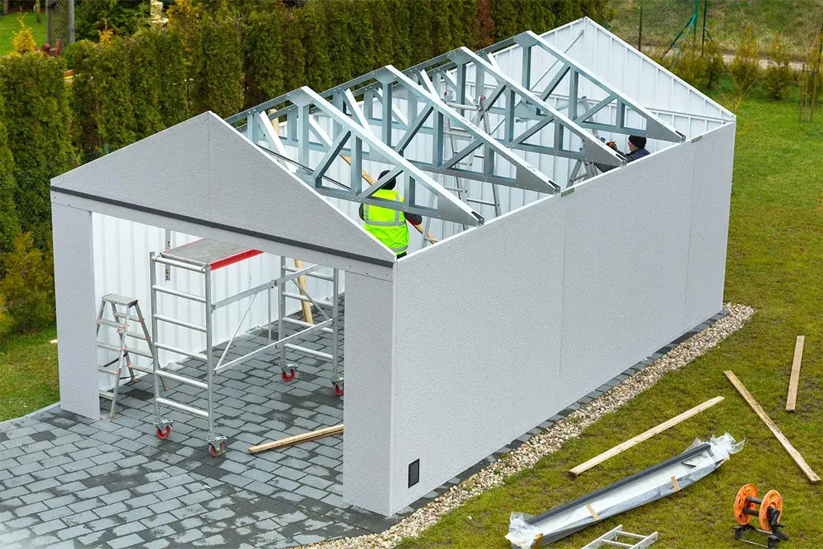 Budowa garażu blaszanego w toku. Pracownicy montują konstrukcję dachu. W tle widać materiały budowlane.