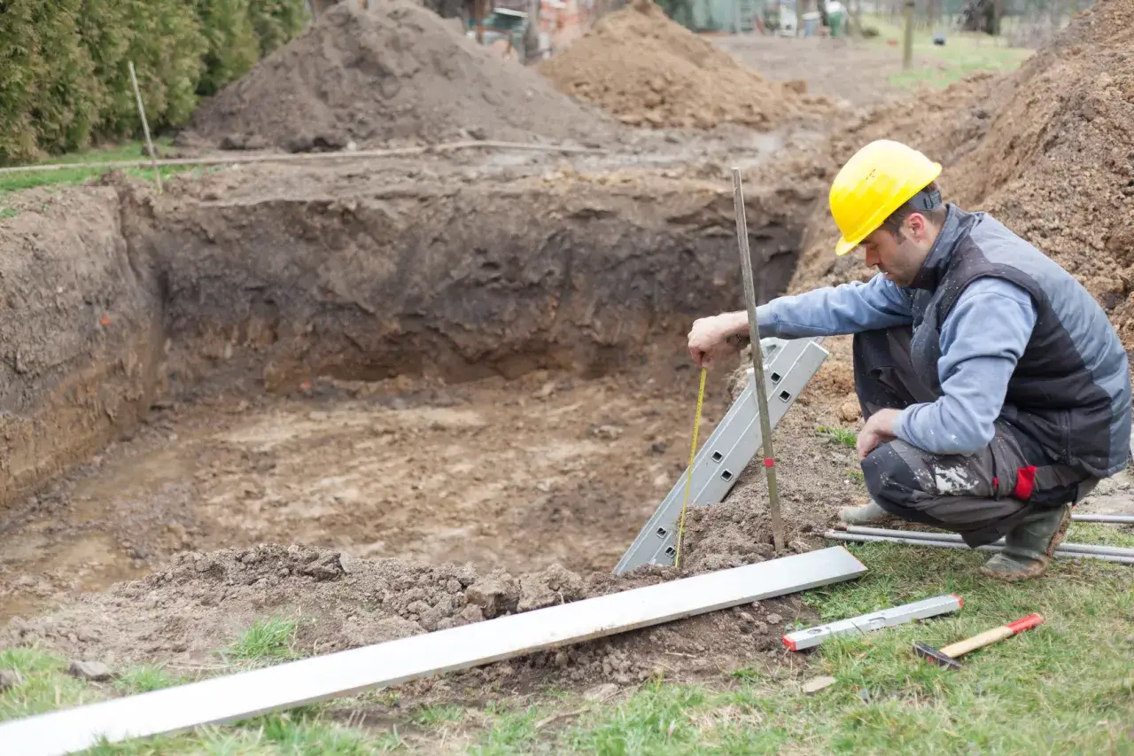 Robotnik w żółtym kasku mierzy głębokość wykopu pod fundament. Wokół hałdy ziemi i narzędzia budowlane.
