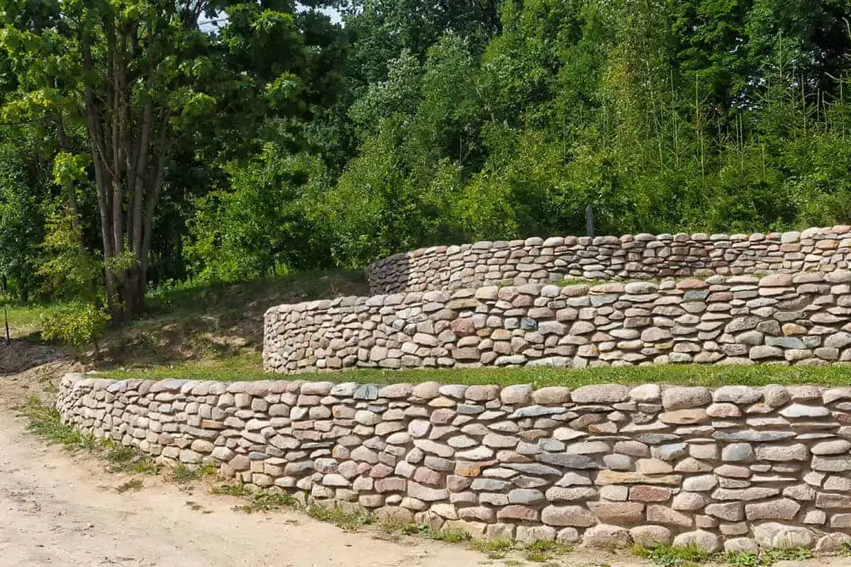 Trzy tarasy z kamienia, tworzące malowniczy mur oporowy, otoczone zielenią i drzewami.