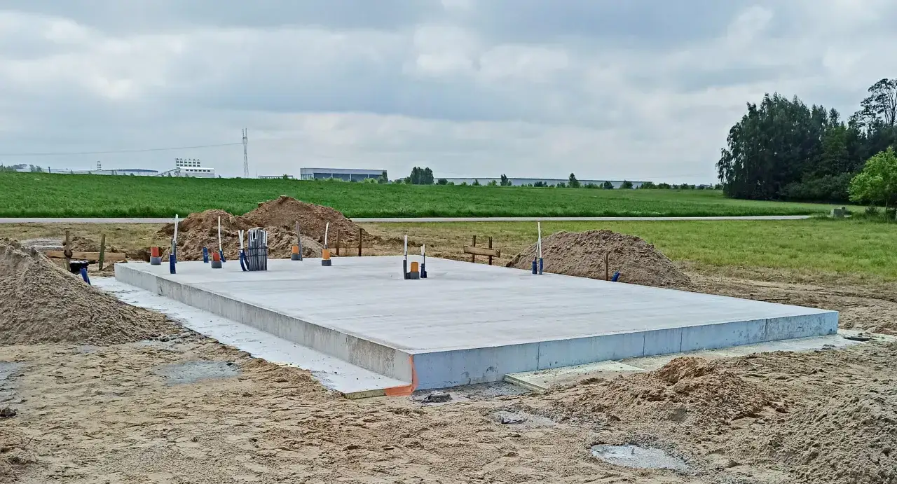 Solidny fundament pod taras gotowy do dalszych prac. Wokół plac budowy z piaskiem i zielone pola w tle.