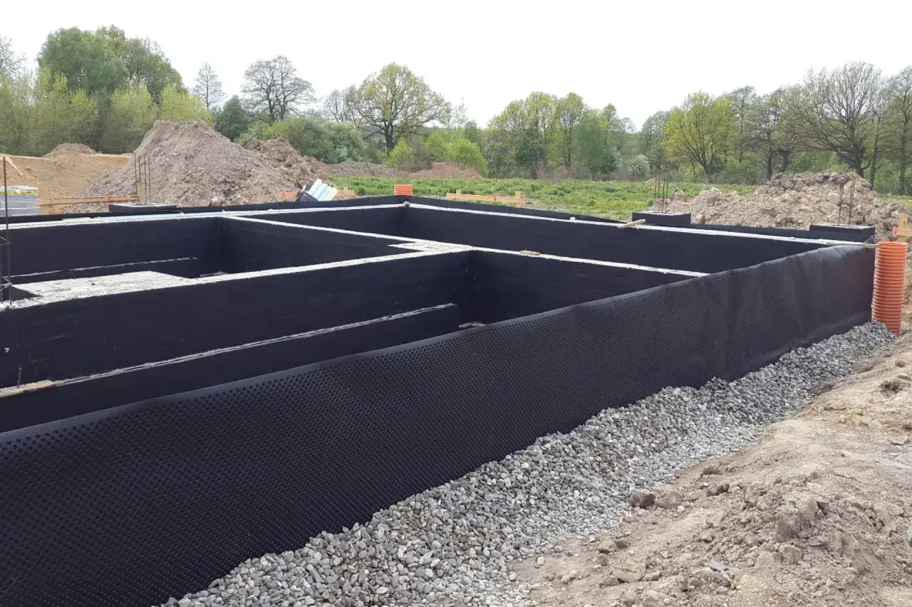 Fundamenty domu w trakcie budowy, z czarną izolacją ścian fundamentowych i otoczone żwirem.
