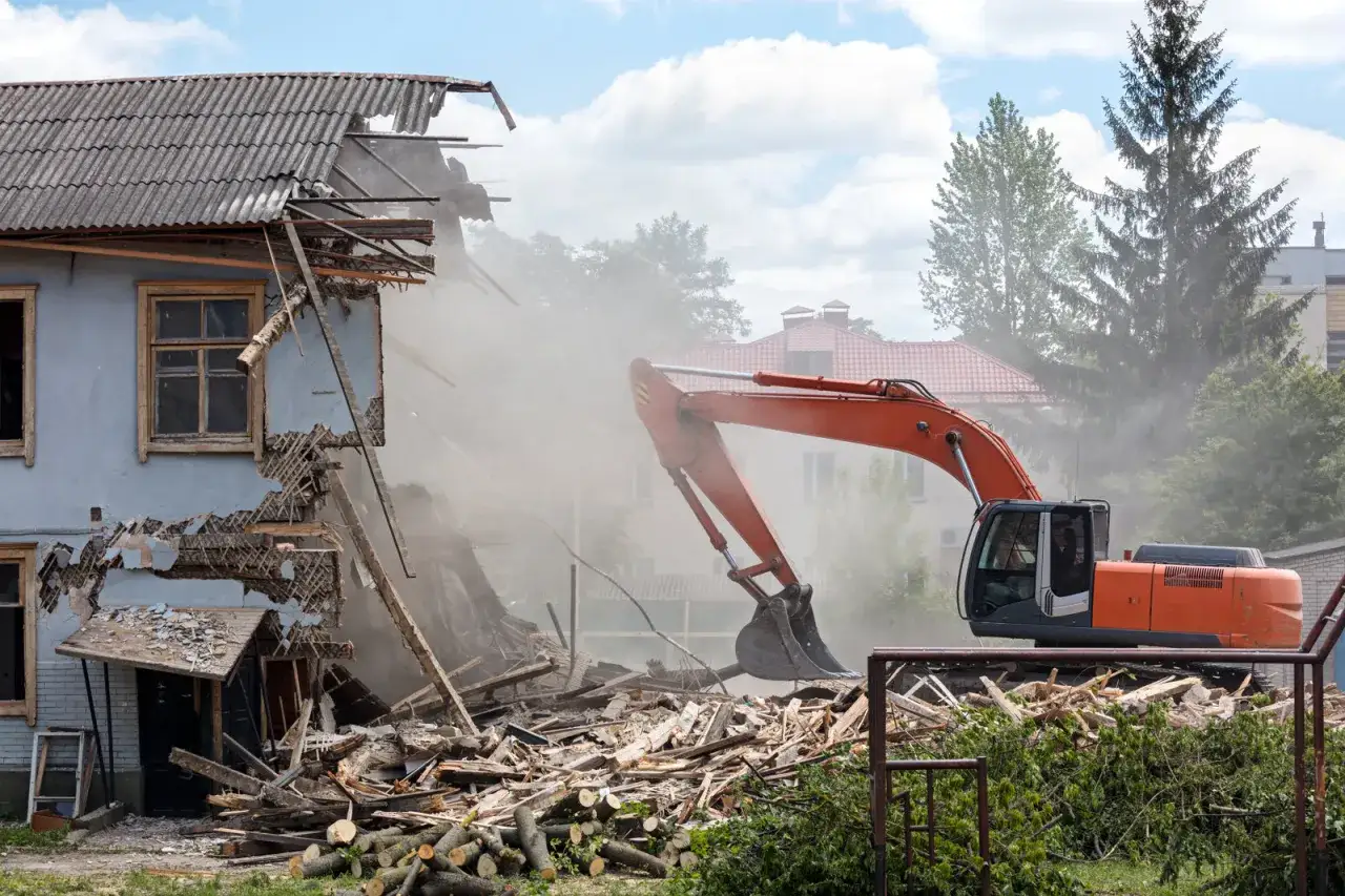 Koparka rozbiera stary dom, unosząc tumany kurzu. Widać zniszczoną konstrukcję i stertę gruzu.