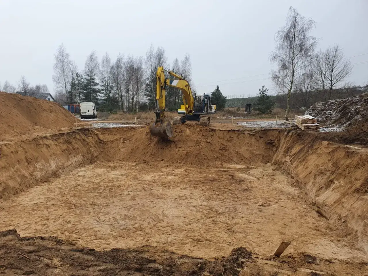 Koparka pracuje przy wykopie pod ławy fundamentowe. W tle widać drzewa i budynki.
