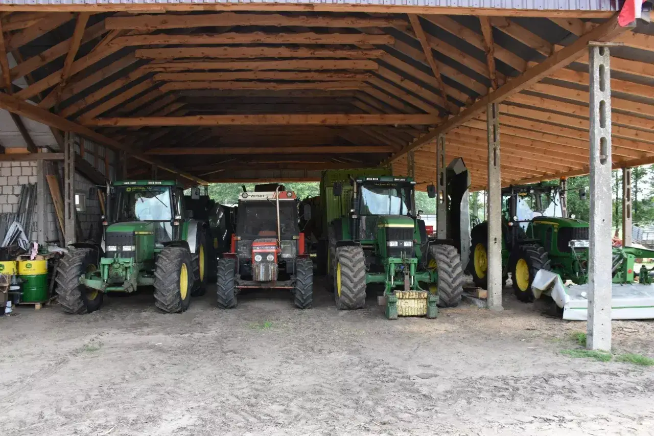 Cztery traktory stoją pod drewnianą wiatą stalową rolniczą. Dwa zielone John Deere, jeden czerwony Zetor i jeden zielony z przyczepą.